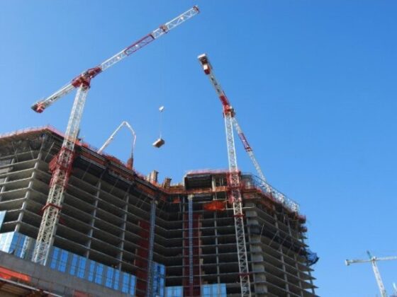 Two red-and-white construction cranes over a partially built high-rise with exposed concrete floors against a clear blue sky.