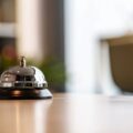 Chrome hotel reception bell on a dark base resting on a wooden counter with a blurred lobby background. Frequented by front desk staff to call for attention.