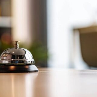 Chrome hotel reception bell on a dark base resting on a wooden counter with a blurred lobby background. Frequented by front desk staff to call for attention.