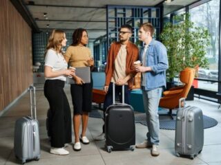 Four travelers with rolling suitcases chat in a modern hotel lobby, one holding a laptop and another with a coffee cup.