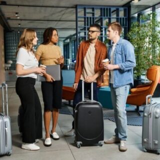 Four travelers with rolling suitcases chat in a modern hotel lobby, one holding a laptop and another with a coffee cup.