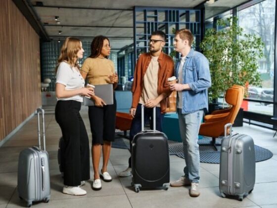 Four travelers with rolling suitcases chat in a modern hotel lobby, one holding a laptop and another with a coffee cup.