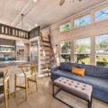 Open-plan loft living area with a blue sofa, tufted ottoman, wooden ladder to a mezzanine, and bright kitchen. Large windows bring in natural light.