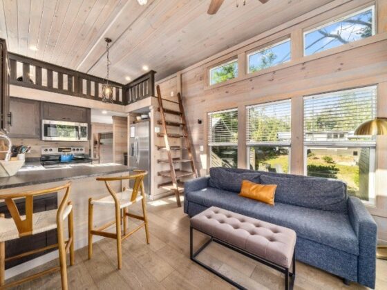 Open-plan loft living area with a blue sofa, tufted ottoman, wooden ladder to a mezzanine, and bright kitchen. Large windows bring in natural light.