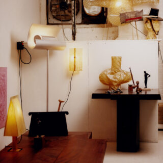 An art studio corner with white cabinetry, abstract sculptures, and hanging lamps under warm lighting.