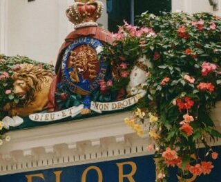 Floral storefront with colorful hanging baskets surrounding a crest and lion statue above the sign that reads 'FLOR' (florist).”,