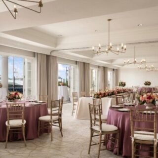 Elegant banquet hall set for a reception with round tables in purple linens, gold chairs, and floral centerpieces under chandeliers.