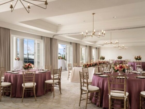 Elegant banquet hall set for a reception with round tables in purple linens, gold chairs, and floral centerpieces under chandeliers.