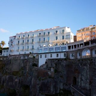 Cliffside white hotel buildings overlooking a rocky sea wall under a clear blue sky with palm trees nearby