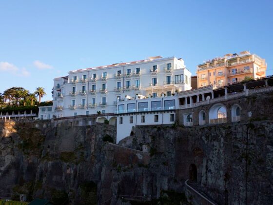 Cliffside white hotel buildings overlooking a rocky sea wall under a clear blue sky with palm trees nearby
