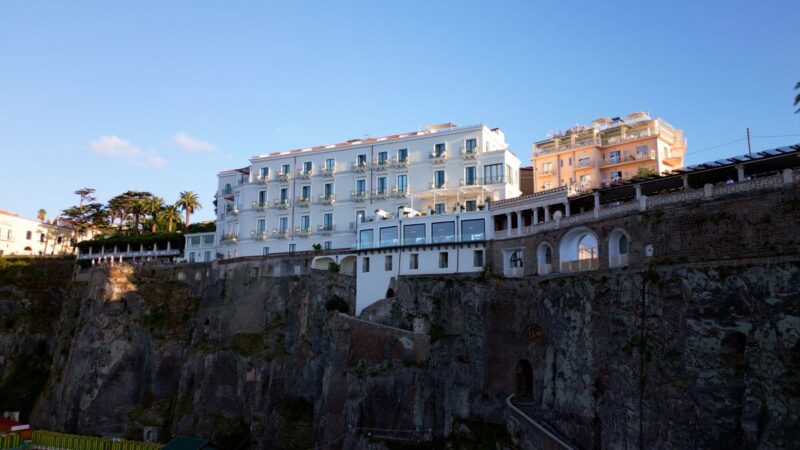 Cliffside white hotel buildings overlooking a rocky sea wall under a clear blue sky with palm trees nearby