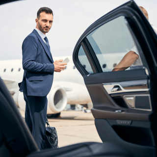 A man in a blue suit stands on an airport tarmac beside an open car door, holding a phone with a briefcase on the ground, near a white jet-shaped aircraft in the background.