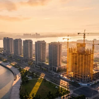 Aerial view of a coastal city at sunset with tall buildings along the water and construction cranes in the foreground.