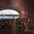 Speaker at a podium addressing a large audience in a curved, brown-walled auditorium with a wide arched window showing a river and city skyline.