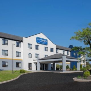 White multi-story hotel with a blue 'City Express by Marriott' sign above a covered entrance, curved driveway, and surrounding greenery under a clear blue sky.