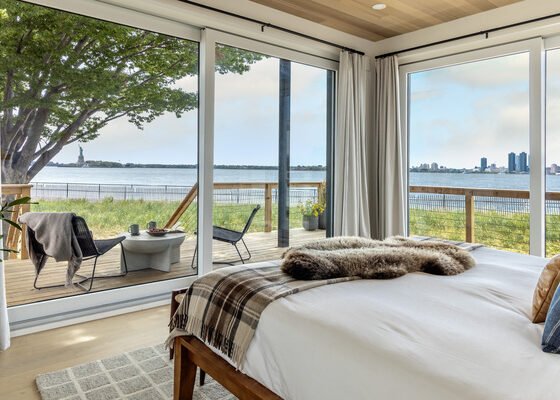 Bedroom with floor-to-ceiling glass doors opening to a wooden deck by the water; a fur throw on the bed and outdoor seating.