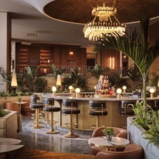 Elegant hotel bar with a curved brass counter, bar stools, warm lighting, and tropical plants in the lounge area.