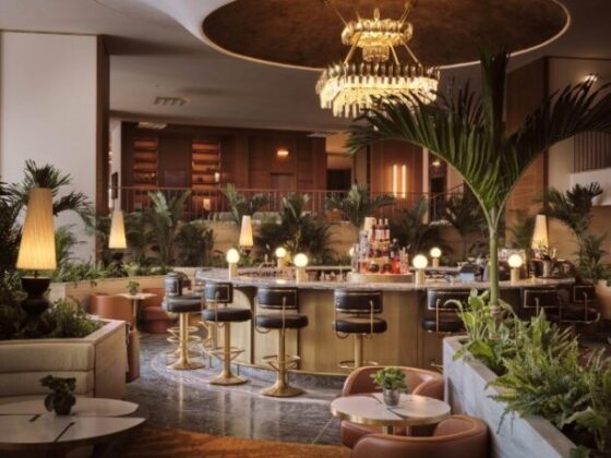 Elegant hotel bar with a curved brass counter, bar stools, warm lighting, and tropical plants in the lounge area.