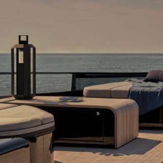 Luxury outdoor yacht deck with beige cushions, blue throw, and a tall black lantern, overlooking the sea at sunset.