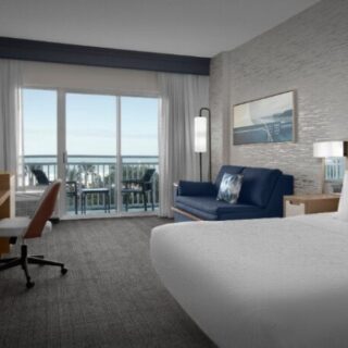 Hotel room with a large white bed, blue sofa, desk, and balcony overlooking an ocean view; bright seating area and modern decor.
