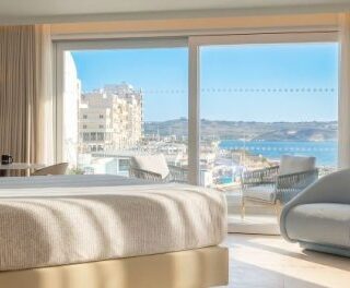 Hotel room with a large bed facing a balcony and ocean view through glass doors.