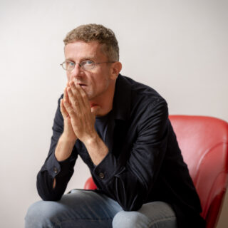 Man with round glasses sits on a red chair, leaning forward with hands clasped near his mouth in a thoughtful pose.