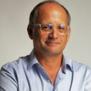 Portrait of a middle‑aged man with glasses wearing a light blue shirt, smiling subtly against a neutral backdrop.