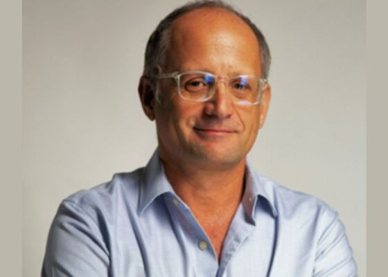 Portrait of a middle‑aged man with glasses wearing a light blue shirt, smiling subtly against a neutral backdrop.