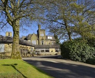 Historic stone building with a tower, set back from a curved driveway, framed by trees and shrubs on a sunny day.