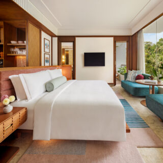 Hotel suite bedroom with a king bed, wooden built‑ins, wall TV, and a teal lounge area by floor‑to‑ceiling windows overlooking lush greenery.
