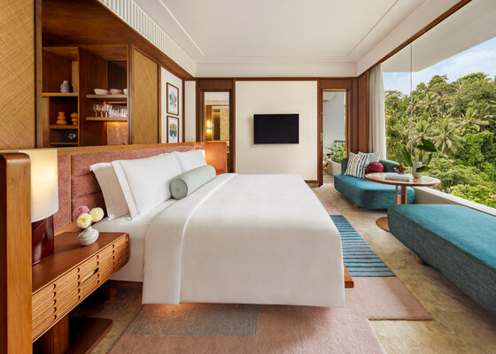 Hotel suite bedroom with a king bed, wooden built‑ins, wall TV, and a teal lounge area by floor‑to‑ceiling windows overlooking lush greenery.