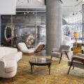 Modern hotel showroom lobby with white curved sofas, wooden lounge chairs, and glass coffee tables on a warm patterned rug; a large blue wall with white text is on the left side.