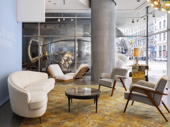 Modern hotel showroom lobby with white curved sofas, wooden lounge chairs, and glass coffee tables on a warm patterned rug; a large blue wall with white text is on the left side.
