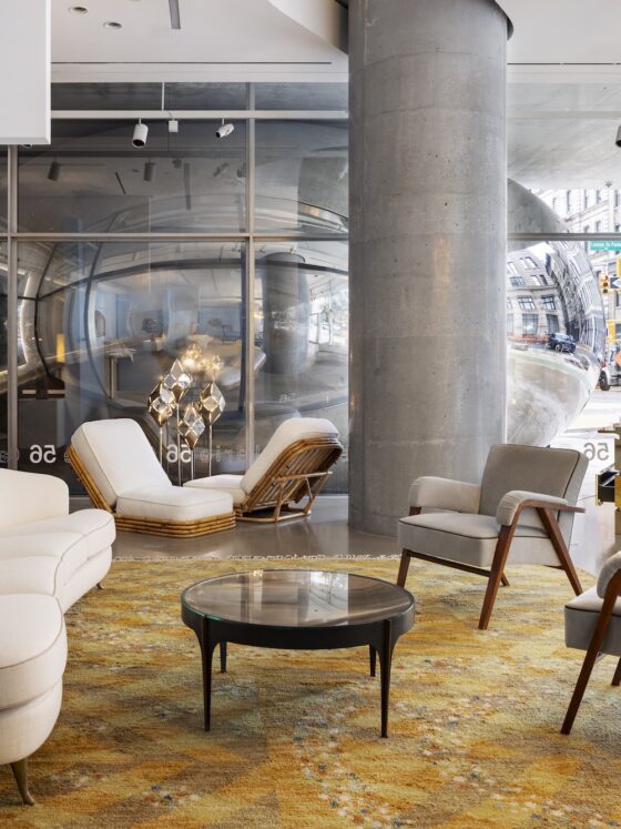 Modern hotel showroom lobby with white curved sofas, wooden lounge chairs, and glass coffee tables on a warm patterned rug; a large blue wall with white text is on the left side.