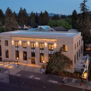 Aerial view of a beige brick municipal building with tall columns and glowing windows on a tree-lined street at dusk, with trees and hills in the background.