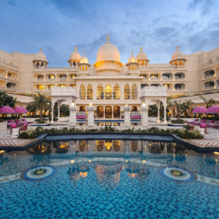 Grand white palace hotel with domes overlooking a blue tiled pool and pink umbrellas in a resort courtyard