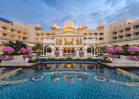 Grand white palace hotel with domes overlooking a blue tiled pool and pink umbrellas in a resort courtyard