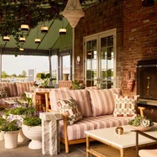Cozy outdoor living area on a patio with a striped sofa, wicker chairs, and a brick fireplace. Plants line the space under hanging lanterns.