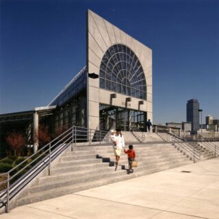 Exterior view of a modern building with a large circular arched glass facade and wide concrete steps, pedestrians walking down the stairs against a blue sky.