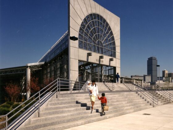 Exterior view of a modern building with a large circular arched glass facade and wide concrete steps, pedestrians walking down the stairs against a blue sky.