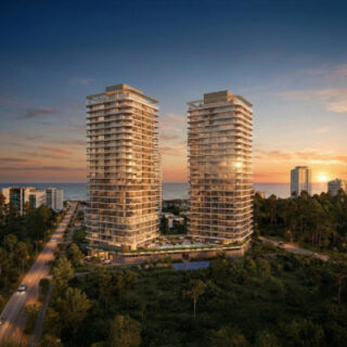 Twin glass high-rise residential towers rise above a green, tree-filled coastline at sunset, with the sea on the horizon and smaller buildings nearby.