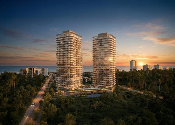 Twin glass high-rise residential towers rise above a green, tree-filled coastline at sunset, with the sea on the horizon and smaller buildings nearby.