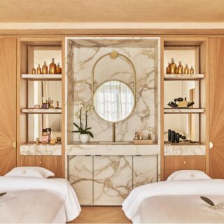 Two single beds with white linens face a marble vanity with a round mirror and gold frame in a wood-paneled room filled with cosmetic bottles on open shelves.