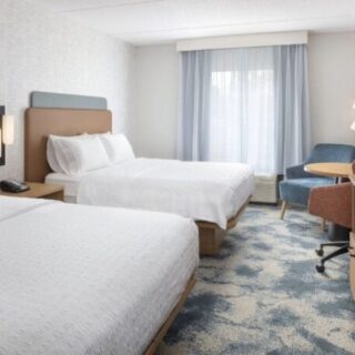 Hotel room with two neatly made beds, nightstands, and wall lamps; window with sheer curtains in the background.
