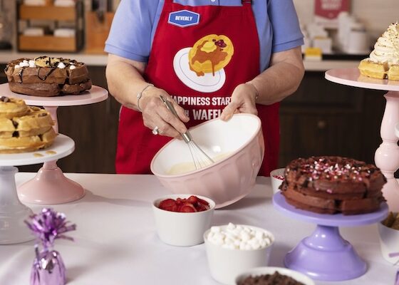 Bakery worker in a red apron whisking batter in a bowl at a dessert table with cakes on pedestals nearby.