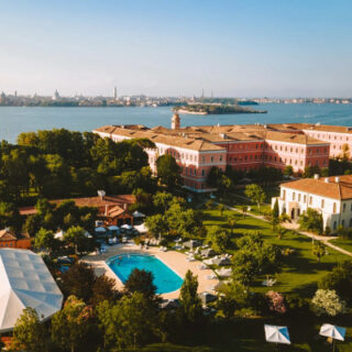 Aerial view of a waterfront estate with red-brick buildings, lush gardens, a large swimming pool, and white tents.