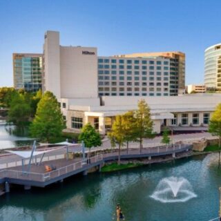 Hilton hotel complex along a river with a pedestrian boardwalk and fountain in the water ahead.
