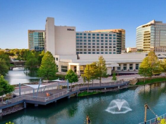 Hilton hotel complex along a river with a pedestrian boardwalk and fountain in the water ahead.