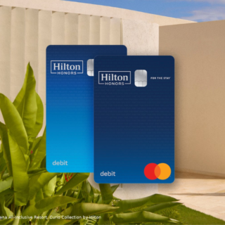 Two blue Hilton Honors debit cards over a tropical resort scene; a woman walks along a beige wall beside plants.
