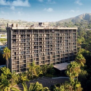 A large multi-story hotel named Hotel La Jolla sits among palm trees, with a hillside landscape visible in the background and an aerial view from above.